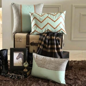 May include: Three decorative cushions in mint green and textured grey fabric. One square cushion has a central mint panel, one rectangular cushion features a brown chevron pattern, and one rectangular cushion has a split design of mint and grey fabric.