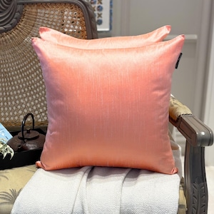 May include: A square throw cushion in a solid peach colour with a subtle vertical slub texture. The cushion features a smooth, slightly lustrous finish and a small black fabric tag attached to the right side seam, presenting a clean, uniform appearance.