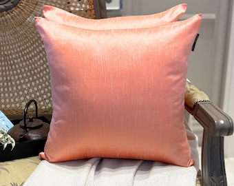 May include: A square throw pillow in a solid peach-colored fabric with a subtle vertical slub texture. The pillow features a smooth, slightly lustrous finish and a small black brand tag attached to the upper right side seam.