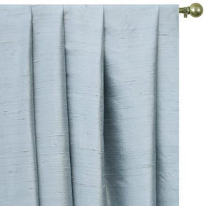May include: A light blue curtain panel with a subtle horizontal slub texture and vertical pleats, suspended from a metallic gold curtain rod featuring a spherical finial. The fabric has a slight sheen and a consistent, cool-toned color throughout.