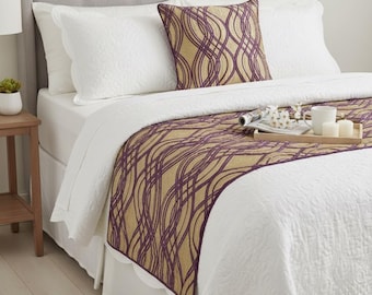 May include: A bed featuring a white quilted coverlet with a scalloped edge, a gold and purple wavy-patterned bed runner, and a matching square throw pillow. The bed has a light gray tufted headboard and white pillows.