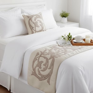 Puede incluir: Un juego de cama blanco que incluye un camino de cama beige y un cojín cuadrado a juego, ambos decorados con un patrón de volutas beige bordado. La cama está vestida con sábanas blancas lisas, con una bandeja de madera con una taza y velas.