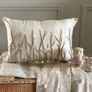 Op de afbeelding: Een rechthoekig crèmekleurig sierkussen met een geborduurd patroon van verticale beige koordlijnen, afgewerkt met kleine witte ronde knopen. Naast het kussen staan twee kleine, lichtgekleurde uitgesneden stenen figuren met schelpaccenten.