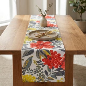 May include: A rectangular table runner featuring a vibrant floral pattern with large red and yellow blossoms, dark gray foliage, and green leaves on a white background. A round beige ceramic vase and a plate with gold cutlery sit on the runner.