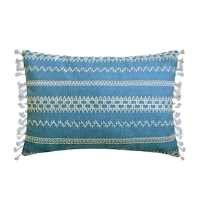 Op de afbeelding: Een blauw en wit gepatroneerd kussen met witte franjes. Het kussen heeft een geometrisch ontwerp met witte stiksels.