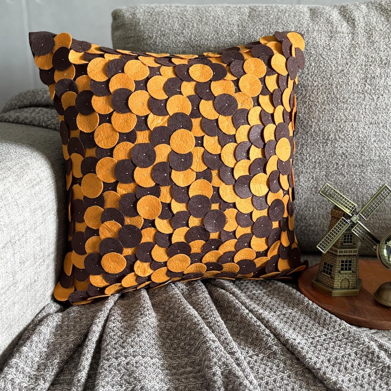 Pode incluir: Almofada decorativa quadrada com um padr&atilde;o de c&iacute;rculos castanhos e laranja. A almofada est&aacute; colocada num sof&aacute; cinzento e numa manta cinzenta. A almofada &eacute; um artigo de decora&ccedil;&atilde;o para casa.