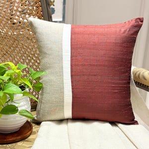 Può includere: Un cuscino quadrato con un design a blocchi di colore, caratterizzato da una sezione beige chiaro testurizzata a sinistra, una sottile striscia verticale bianca al centro e una sezione rosso bordeaux intenso sulla destra in tessuto fiammato.