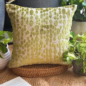 May include: Square cushion featuring a pattern of lime green circles on a light beige backdrop. The cushion is placed on a round wicker base, suggesting a home decor theme. The cushion measures approximately 40cm x 40cm.