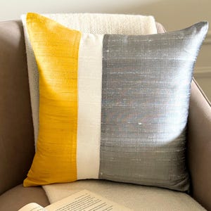 Op de afbeelding: Een vierkant sierkussen met een kleurblokontwerp met een verticaal mosterdgeel paneel aan de linkerkant, een smalle crèmekleurige middenstreep en een breed metallic zilvergrijs paneel aan de rechterkant, alles met een zichtbare horizontale slubtextuur.