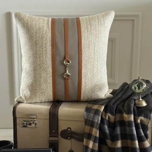 May include: A square throw cushion with a textured beige fabric cover featuring a central vertical grey panel flanked by two brown leather-like straps. A gold-toned metal horsebit hardware detail is centred on the grey panel.