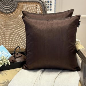 May include: Two square, dark brown throw pillows with a subtle sheen, resting on a light-colored blanket. The pillows are on a wicker chair, with a decorative teapot and a book visible in the background. The pillows add a touch of elegance to the decor.
