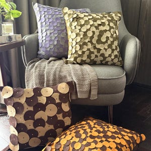 Pode incluir: Quatro almofadas decorativas com um padr&atilde;o circular em tons de roxo, verde-oliva, castanho e laranja. As almofadas est&atilde;o dispostas numa poltrona cinzenta com uma manta de cor neutra.
