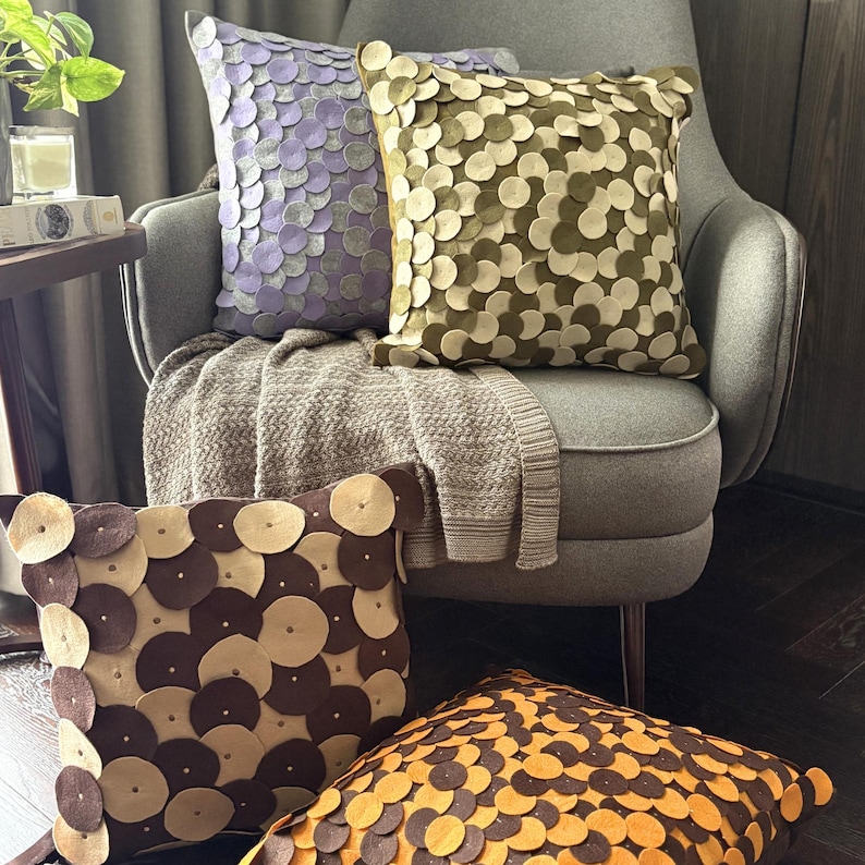 Pode incluir: Quatro almofadas decorativas com um padr&atilde;o circular em tons de roxo, verde-oliva, castanho e laranja. As almofadas est&atilde;o dispostas numa poltrona cinzenta com uma manta de cor neutra.