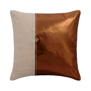 May include: A square throw pillow featuring a two-tone design. The left side is made of a light beige, textured fabric, while the right side is a smooth, metallic copper-colored material. A single vertical white stitch separates the two panels.