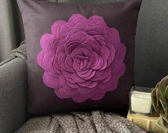 May include: A square throw pillow in a deep plum color featuring a large, centered, three-dimensional flower applique. The flower is constructed from multiple layers of felt-like fabric petals in a vibrant magenta shade, creating a full, rounded bloom.