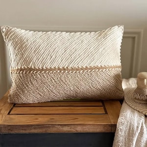 May include: A rectangular throw pillow featuring a two-tone design with an off-white upper section and a tan lower section. Both sections display a diagonal corded texture, separated by a horizontal braided trim across the centre of the pillow.