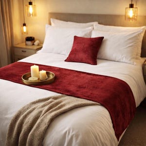 Puede incluir: Una cama con ropa de cama blanca, un camino de cama de terciopelo rojo intenso y un cojín cuadrado a juego. Una bandeja circular dorada sobre el camino sostiene tres velas blancas de diferentes alturas, iluminadas por lámparas colgantes cálidas.