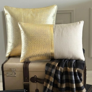 Puede incluir: Dos cojines decorativos. Un cojín cuadrado presenta un acabado dorado metálico con una textura de cuadrícula. El segundo cojín rectangular está dividido entre un lado dorado metálico con cuadrícula y un lado de tela beige tipo lino.