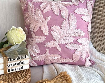 May include: A square throw pillow in a dusty mauve color featuring a textured, raised leaf pattern in a lighter pink shade. The pillow is positioned next to a stack of three white decorative blocks with the words Thankful, Grateful, Blessed.