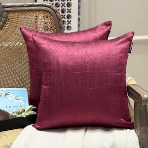 Può includere: Due cuscini color magenta con superficie strutturata sono esposti su una sedia. I cuscini hanno una forma quadrata e sono realizzati in un tessuto lucido. I cuscini sono posizionati su un cuscino di colore chiaro.