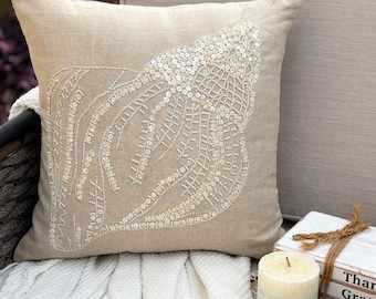 May include: A square beige throw pillow featuring a large, detailed seashell design crafted from white and cream-colored beads and sequins. The intricate shell pattern is centered on the front of the fabric, showcasing a textured, shimmering finish.