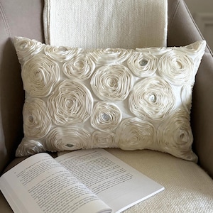 May include: A rectangular off-white throw pillow featuring a dense surface texture of layered fabric rosettes. Each rosette is constructed from coiled ribbon-like material, with small, clear, circular buttons centered within several of the floral shapes.