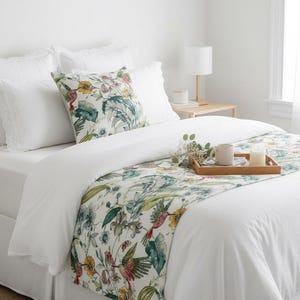 May include: A white bed featuring a decorative runner and a square throw pillow, both adorned with a vibrant botanical print of hummingbirds, tropical leaves, and yellow flowers. A wooden tray holds a white mug and a candle on the bed.