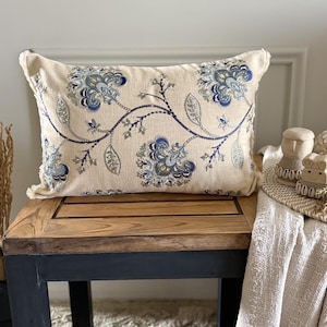 May include: A rectangular throw cushion with a cream-coloured base featuring an intricate embroidered floral pattern in shades of navy blue, light blue, and silver. The cushion has a frayed edge finish and a central vine design connecting the flowers.