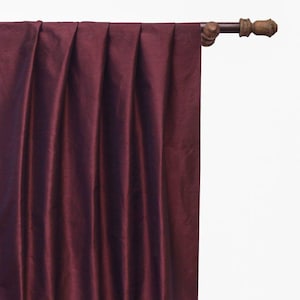 May include: A deep burgundy curtain panel with a subtle sheen and vertical pleats, suspended from a dark brown wooden curtain rod featuring decorative finials. The fabric displays a smooth texture with soft folds cascading downward from the rod.