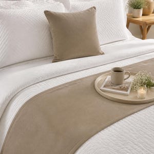 Puede incluir: Una cama con ropa de cama blanca acolchada, un cojín cuadrado de color tostado liso y un camino de cama a juego. Sobre el camino hay una bandeja redonda con una taza beige acanalada, un libro pequeño, una rama de paniculata y una vela.