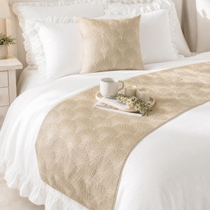 Puede incluir: Un cojín cuadrado y un camino de cama rectangular largo con un patrón de abanico en dorado y blanco. El juego de cama se muestra sobre una cama blanca con almohadas blancas con volantes y una funda nórdica blanca, con una taza y velas.