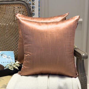 Puede incluir: Dos cojines cuadrados decorativos de color rosa con un sutil estampado de rayas verticales. Los cojines están hechos de una tela brillante y satinada y están colocados sobre una silla de color claro. Los cojines añaden un toque de elegancia a cualquier espacio habitable.