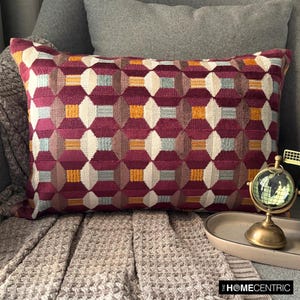 Könnte beinhalten: Ein burgunderfarbenes Kissen mit einem geometrischen Muster in Orange-, Weiß- und Blautönen. Das Kissen liegt auf einem grauen Sofa mit einer braunen Strickdecke.