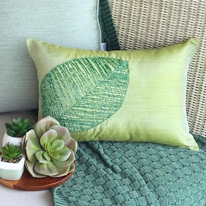 Puede incluir: Un cojín rectangular de color verde claro que presenta un gran motivo de hoja central adornado con intrincados abalorios verdes. El cojín tiene una textura horizontal sutil y se coloca cerca de una planta suculenta y una manta verde texturizada.