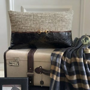 Puede incluir: Un cojín rectangular con una sección superior de tejido texturizado en beige y gris y una sección inferior negra lisa, separadas por un borde marrón fino con un detalle de herraje metálico dorado en el centro.