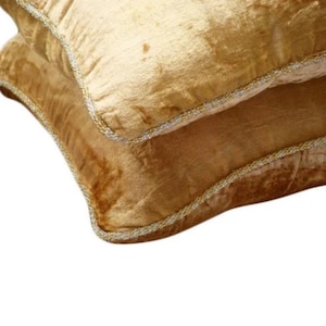 Può includere: Una coppia di cuscini quadrati rivestiti in tessuto di velluto giallo dorato. Ogni cuscino presenta una distinta consistenza scintillante ed è rifinito con un bordo decorativo in cordoncino metallico argentato lungo i lati, creando un profilo definito.