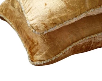 May include: A pair of square throw pillows covered in a golden yellow velvet fabric. Each pillow features a distinct, shimmering texture and is finished with a decorative silver metallic cord trim along the edges, creating a defined border.