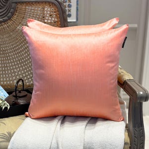 May include: Two square peach-coloured throw cushions with a textured, silky appearance. The cushions are placed on a chair with a light-coloured blanket. The cushions have a subtle sheen and a classic design, suitable for home decor.