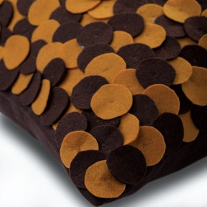Pode incluir: Uma almofada quadrada decorativa com base castanha escura, adornada com c&iacute;rculos de feltro sobrepostos em tons de castanho e amarelo mostarda. O design da almofada cria um efeito texturizado, adequado para decora&ccedil;&atilde;o de casa.