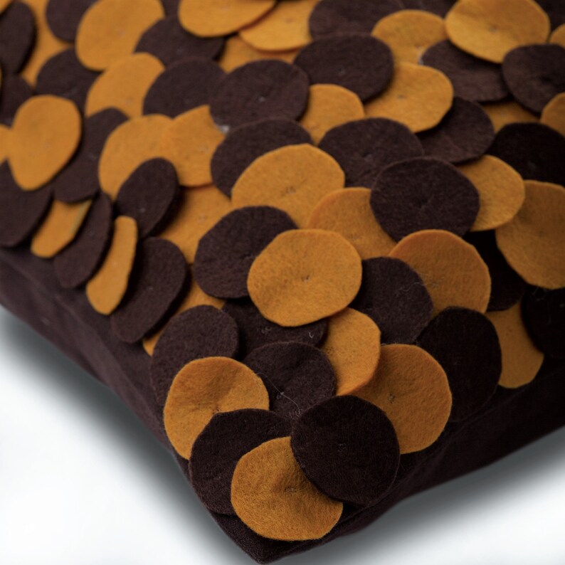 Pode incluir: Uma almofada quadrada decorativa com base castanha escura, adornada com c&iacute;rculos de feltro sobrepostos em tons de castanho e amarelo mostarda. O design da almofada cria um efeito texturizado, adequado para decora&ccedil;&atilde;o de casa.