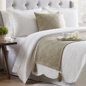 May include: A white quilted bedspread with scalloped edges, two matching white quilted pillow shams, a square beige accent pillow with a repeating scroll pattern, and a long beige bed runner featuring the same scroll pattern and tassel ends.