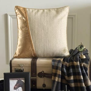 May include: A square throw cushion featuring a split design with a smooth metallic gold panel on the left and a textured cream-coloured fabric on the right. The cushion is positioned atop a vintage-style suitcase with brown leather straps.