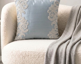 May include: A square throw pillow in a light blue color featuring intricate, cream-colored embroidered scrollwork patterns along the left and right vertical edges. The pillow has a smooth, slightly lustrous finish and a structured, square shape.