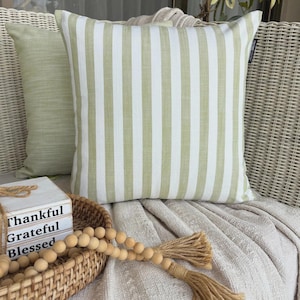 May include: A square throw cushion featuring vertical sage green and white stripes. The cushion has a small black fabric tag on the right side. It is positioned in front of a solid light green cushion and rests on a textured off-white fabric surface.