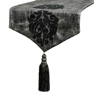 Puede incluir: Un camino de mesa de terciopelo gris oscuro con un intrincado patrón de damasco hecho de cuentas negras centrado cerca del extremo puntiagudo. El camino termina con una borla de tela larga y a juego en gris oscuro en la punta.