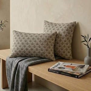 Op de afbeelding: Twee sierkussens met een herhalend beige en bruin bladpatroon. Het ene kussen is rechthoekig en het andere is vierkant. Ze liggen op een houten oppervlak naast een grijze plaid en een stapel tijdschriften met het woord Monocle zichtbaar.