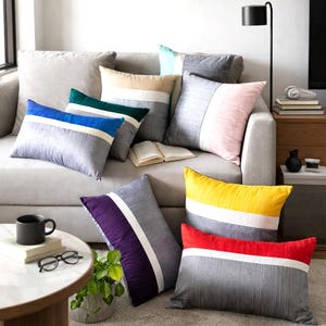 May include: A collection of rectangular and square throw pillows featuring a textured gray base with a prominent horizontal white stripe and a secondary block of color in blue, green, beige, pink, yellow, purple, or red, arranged on a sofa.