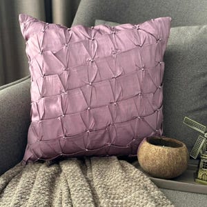 Può includere: Un cuscino quadrato color malva polveroso caratterizzato da un design in tessuto arricciato e pieghettato su tutta la superficie anteriore. Il materiale presenta una lucentezza sottile ed è accentuato da piccoli dettagli a pieghe che formano una griglia.