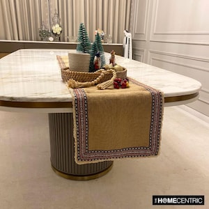 May include: A rectangular tan burlap table runner featuring a decorative border with a black and red geometric cross-stitch pattern and frayed edges. The runner is draped over a white marble table with a round, fluted base and gold trim.