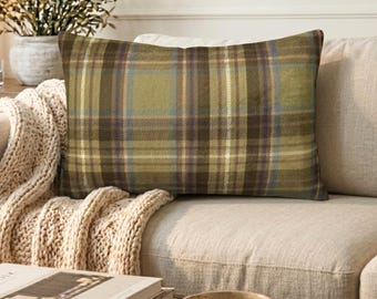 May include: A rectangular throw pillow featuring a plaid pattern in shades of olive green, dark brown, and cream. The fabric displays a woven texture with intersecting horizontal and vertical lines forming a classic grid design across the surface.